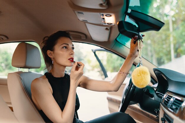 Jeune femme élégante à la recherche dans le miroir de vue de voiture tout en appliquant