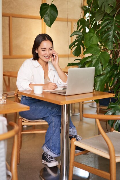 Une jeune femme élégante répond à un appel téléphonique assis dans un café avec un ordinateur portable travaillant et parlant sur un smartphone