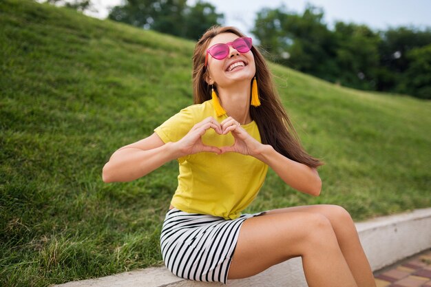 Jeune femme élégante en riant s'amuser dans le parc de la ville, souriant humeur joyeuse, portant haut jaune, mini jupe rayée, lunettes de soleil roses, tendance de la mode de style d'été, montrant le signe du coeur