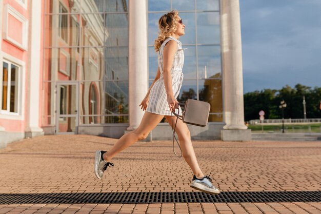 Jeune femme élégante et séduisante en cours d'exécution sautant drôle dans des baskets dans la rue de la ville en robe blanche de style de mode d'été portant des lunettes de soleil et un sac à main