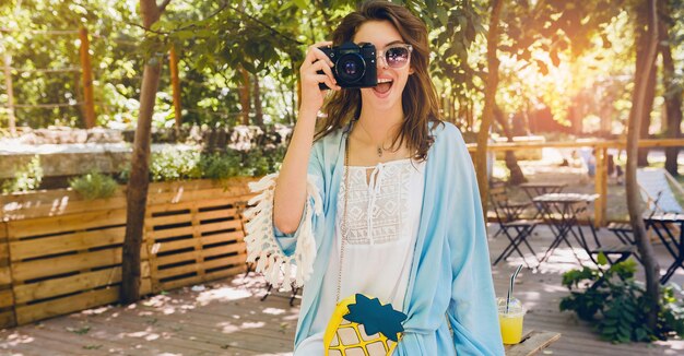 Jeune femme élégante et séduisante dans le parc, style de rue, tendance de la mode estivale, cape bleue, robe boho blanche, accessoires, prendre des photos sur appareil photo vintage, souriant, émotion heureuse, ensoleillée