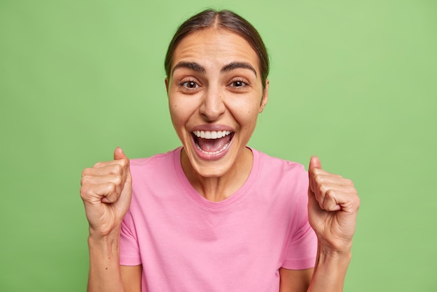 Une jeune femme émotionnelle joyeuse s'exclame joyeusement en serrant les poings avec une expression de visage euphorique, des rires se réjouissent positivement de bonnes nouvelles isolées sur fond vert. Concept de grande victoire et de réussite