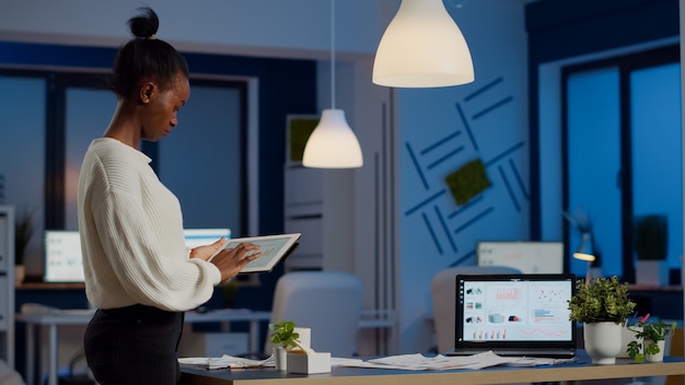 Jeune femme employée debout dans le bureau et analysant les graphiques pendant la pause dans le bureau d'affaires tard dans la nuit