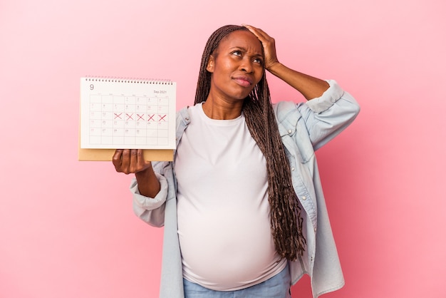 Jeune femme enceinte afro-américaine tenant un calendrier isolé sur fond rose étant choquée, elle s'est souvenue d'une réunion importante.
