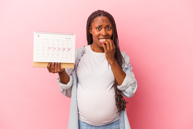 Jeune femme enceinte afro-américaine tenant un calendrier isolé sur fond rose se ronger les ongles, nerveuse et très anxieuse.