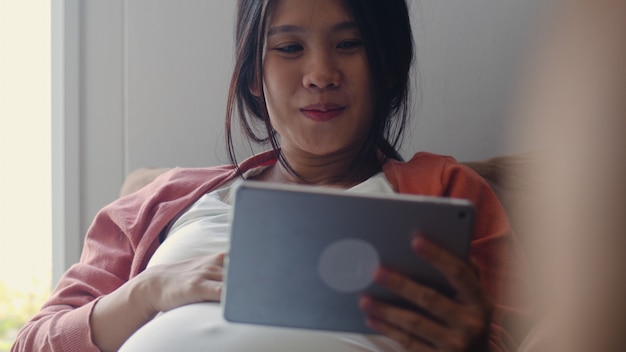 Jeune femme enceinte asiatique à l'aide de la tablette recherche des informations sur la grossesse. Maman se sentant heureuse, souriante, positive et paisible tout en prenant soin de son enfant allongé sur un canapé dans le salon à la maison.