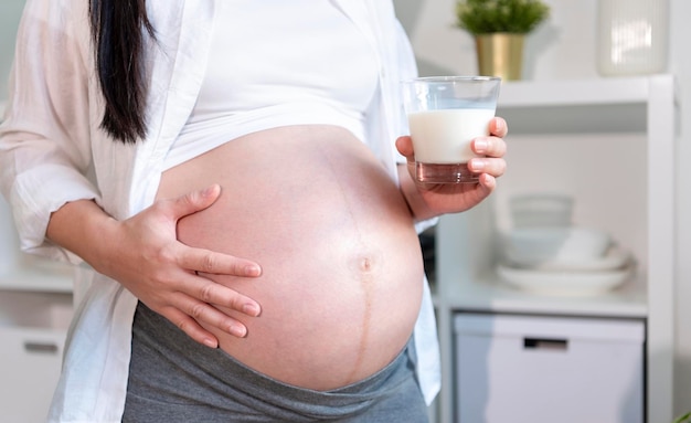 Jeune femme enceinte asiatique debout dans la salle de cuisine tenant un verre de lait frais