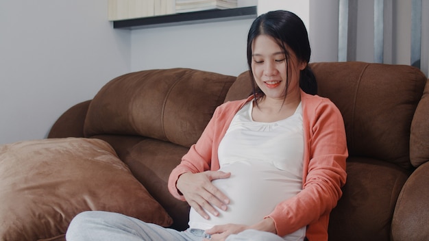 Jeune femme enceinte asiatique tenant son ventre parler avec son enfant. Maman se sentant heureuse, souriante, positive et paisible alors qu’elle prend soin de son bébé, d’une grossesse allongée sur le canapé du salon de la maison