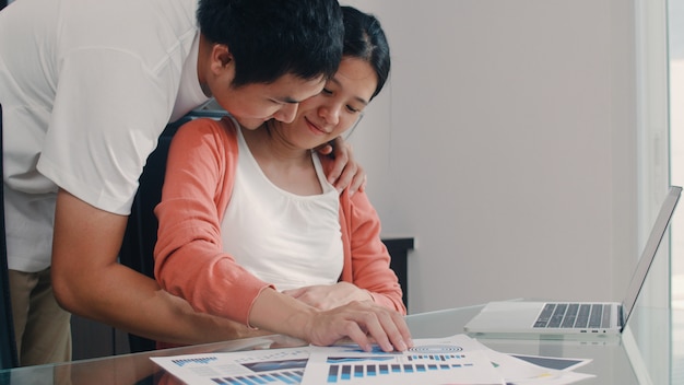 Photo gratuite jeune femme enceinte asiatique utilisant des enregistrements d'ordinateur portable des revenus et des dépenses à la maison. papa touche le ventre de sa femme tout en enregistrant un document financier, budgétaire et financier dans le salon à la maison.