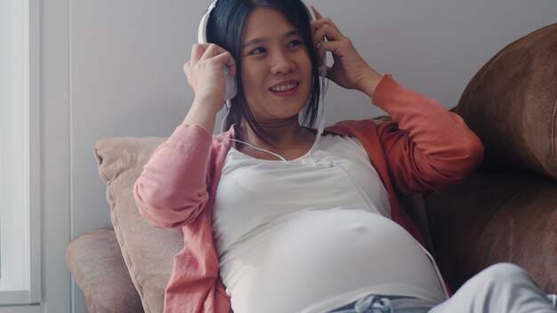 Jeune femme enceinte asiatique utilisant un téléphone et un casque joue de la musique pour bébé dans le ventre. Maman se sentant heureuse, souriante, positive et paisible alors qu’il faut prendre soin de l’enfant couché sur un canapé dans le salon de la maison