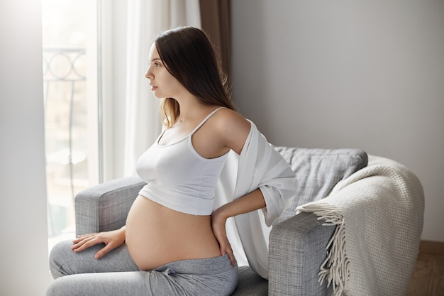 Jeune femme enceinte se frottant le dos pour soulager les douleurs. Rester dans sa maison chaleureuse et confortable.