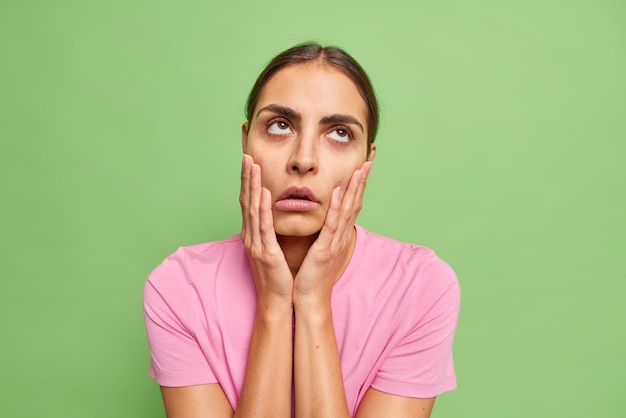 Une jeune femme ennuyée et désintéressée garde les mains sur les joues marre de la conversation terne regarde au-dessus fait facepalm et roule les yeux entend des bêtises porte un t-shirt décontracté isolé sur fond vert.