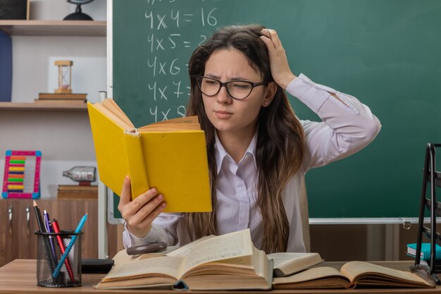 Jeune femme enseignant portant des lunettes avec des livres de lecture étant confus tenant la main sur sa tête assis au bureau de l'école en face du tableau noir en classe