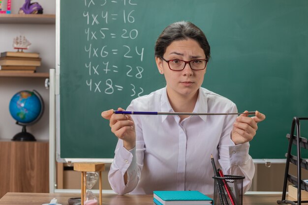 Jeune femme enseignant portant des lunettes regardant à l'avant avec un visage sérieux tenant le pointeur va expliquer la leçon assis au bureau de l'école devant le tableau noir en classe