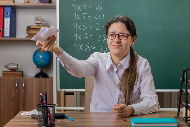 Jeune femme enseignant portant des lunettes tenant des morceaux de papier à la mécontentement assis au bureau de l'école en face du tableau noir en classe