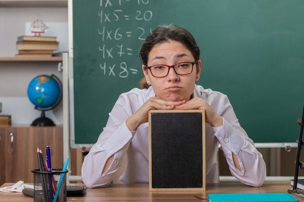 Jeune femme enseignant portant des lunettes tenant petit tableau noir expliquant la leçon à la fatigue et leçon assis au bureau de l'école en face du tableau noir en classe
