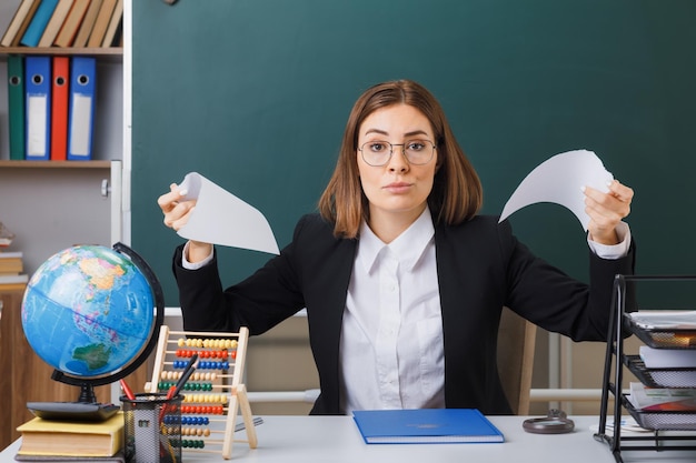 Jeune femme enseignante portant des lunettes assis au bureau de l'école devant le tableau noir dans la salle de classe déchirer un morceau de papier étant mécontent de faire la bouche ironique