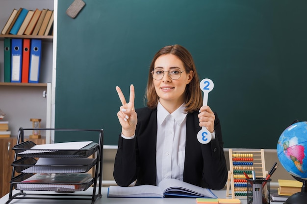 Jeune femme enseignante portant des lunettes assis au bureau de l'école devant le tableau noir dans la salle de classe tenant des plaques d'immatriculation expliquant la leçon montrant vsign souriant joyeusement