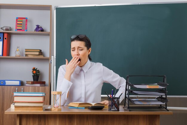 Jeune femme enseignante portant des lunettes assis au bureau de l'école avec un livre devant le tableau noir dans la salle de classe à l'air fatigué et surmené veut dormir en bâillant