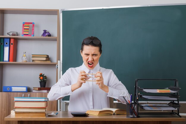 Jeune femme enseignante portant des lunettes assis au bureau de l'école avec un livre devant le tableau noir dans la salle de classe tenant un sablier en colère et frustré criant avec une expression agressive