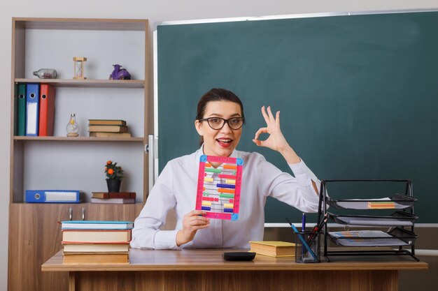 Jeune femme enseignante portant des lunettes tenant un boulier expliquant la leçon souriant amical montrant un signe ok assis au bureau de l'école devant le tableau noir dans la salle de classe
