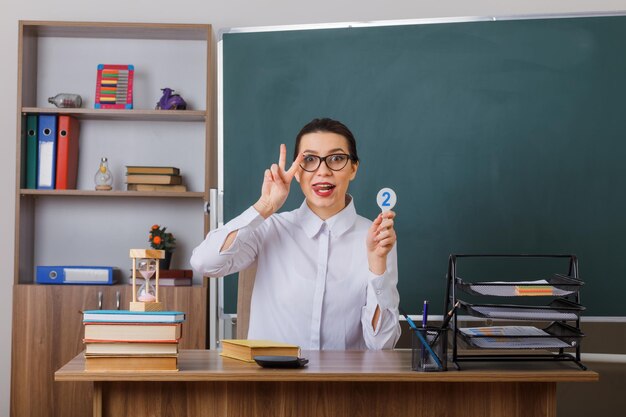 Jeune femme enseignante portant des lunettes tenant la plaque d'immatriculation deux expliquant la leçon montrant vsign qui sort la langue assis au bureau de l'école devant le tableau noir dans la salle de classe