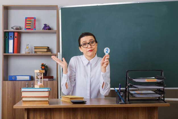 Jeune femme enseignante portant des lunettes tenant la plaque d'immatriculation deux regardant confus levant le bras expliquant la leçon assis au bureau de l'école devant le tableau noir dans la salle de classe