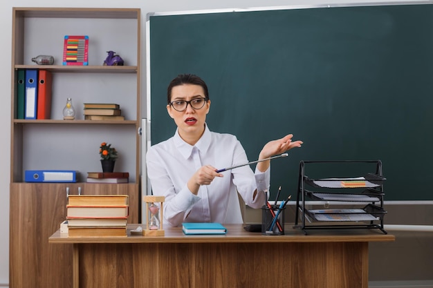 Jeune femme enseignante portant des lunettes tenant un pointeur expliquant la leçon regardant de côté avec une expression sceptique assis au bureau de l'école devant le tableau noir dans la salle de classe