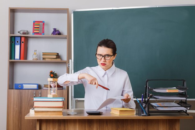 Jeune femme enseignante portant des lunettes vérifiant les devoirs des élèves mécontents et en colère assis au bureau de l'école devant le tableau noir dans la salle de classe