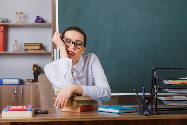 Jeune femme enseignante portant des lunettes vérifiant les devoirs des élèves qui ont l'air fatigué et ennuyé de dormir assis au bureau de l'école devant le tableau noir dans la salle de classe