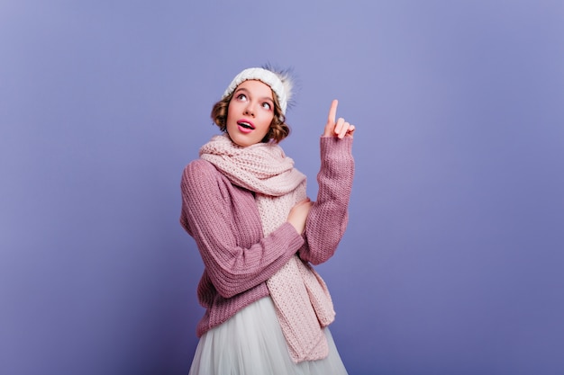 Jeune femme enthousiaste aux cheveux courts posant en longue écharpe. Photo intérieure d'une femme européenne fascinante en tenue d'hiver isolée sur un mur violet.