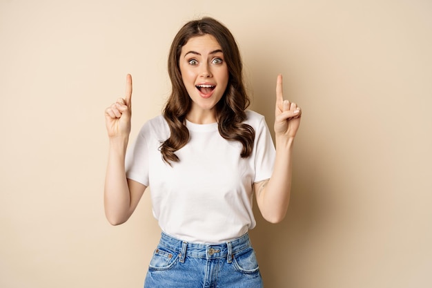 Jeune femme enthousiaste, cliente pointant les doigts vers le haut et souriante, montrant une bannière ou un logo, debout sur fond beige