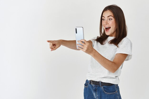 Jeune femme enthousiaste prenant une photo sur un téléphone portable, pointant vers la gauche sur le produit et souriant, enregistrant une vidéo sur un smartphone, mur blanc