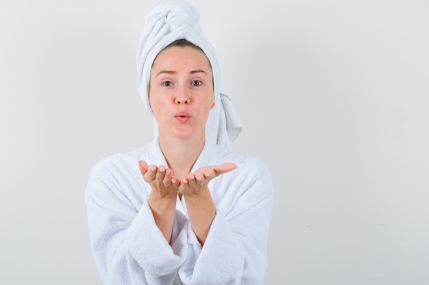 Jeune femme envoyant un baiser d'air avec des lèvres boudeuses en peignoir blanc, serviette et à la jolie vue de face.