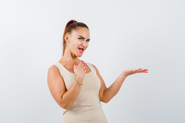 Jeune femme étalant la paume de côté en débardeur beige et à la vue de face, heureux.