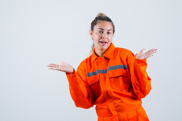 Jeune femme étalant les paumes de côté en uniforme de travailleur et à l'air satisfait. vue de face.