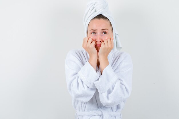 Jeune femme étayant le menton sur les poings en peignoir blanc, serviette et à la pensif, vue de face.