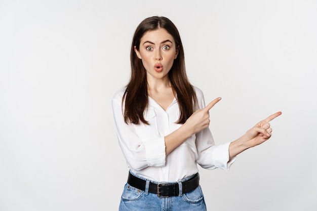 Une jeune femme étonnée pointant les doigts vers le bas, montrant une bannière d'annonce ou de logo, l'air surprise et intriguée, debout sur fond blanc