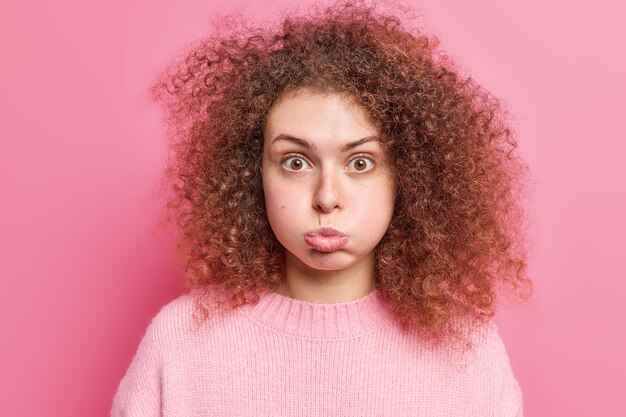 Une jeune femme européenne aux cheveux bouclés offensée souffle des joues fait une grimace vêtue d'un pull décontracté qui retient le souffle et semble étonnamment isolée sur un mur rose. Concept d'expressions de visage