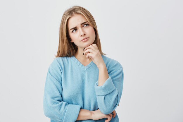 Jeune femme européenne aux yeux ovales et aux cheveux raides, vêtue d'un pull bleu ample, tenant la main sur le menton, regardant avec une expression réfléchie