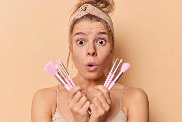 Une jeune femme européenne choquée tient des pinceaux cosmétiques subit une routine de beauté utilise de nouveaux outils réagit à quelque chose d'étonnant porte un bandeau et un t-shirt décontracté isolé sur fond de studio marron