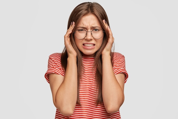 Photo gratuite jeune femme européenne désespérée a de mauvais maux de tête, serre les dents, garde les mains sur la tête, a une expression faciale misérable