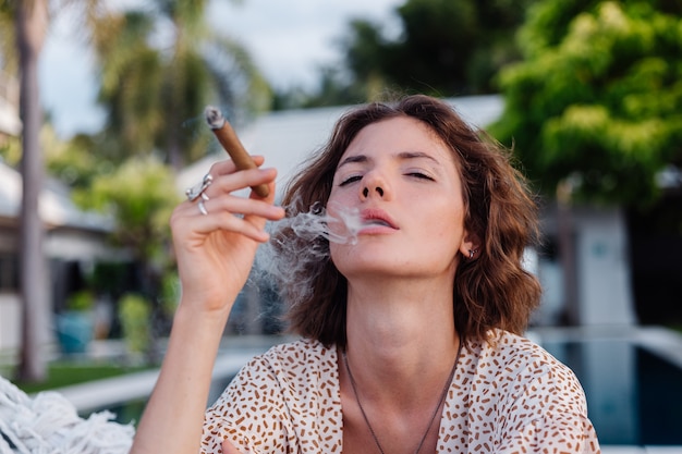 Jeune femme européenne fumant un cigare allongé sur un hamac à l'extérieur de l'hôtel villa de luxe tropical, lumière naturelle du coucher du soleil