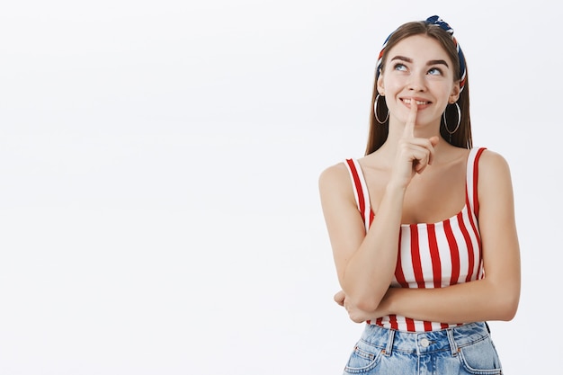 Jeune femme européenne en haut rayé et bandeau souriant largement à la recherche au coin supérieur gauche montrant le geste chut avec l'index sur la bouche