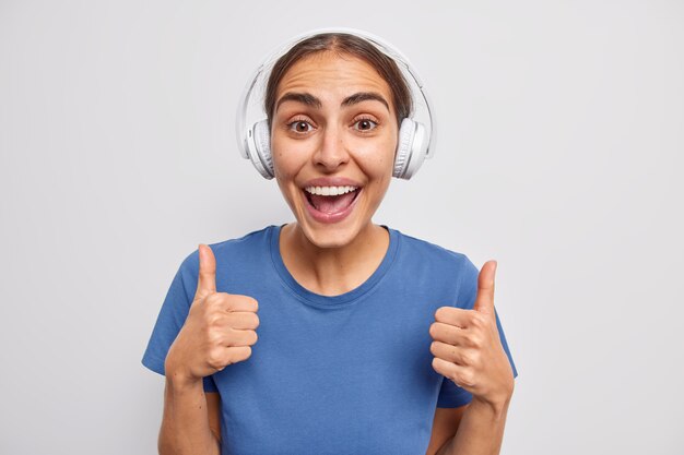 Une jeune femme européenne positive garde les pouces levés est d'accord avec quelque chose qui montre comme un signe être de bonne humeur porte un t-shirt décontracté écoute de la musique dans des écouteurs sans fil pose contre un mur blanc