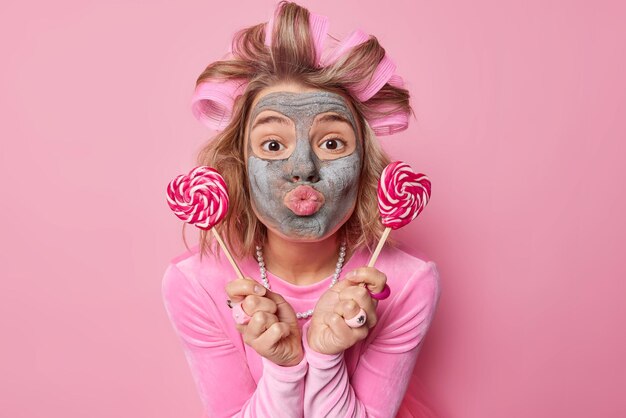 Une jeune femme européenne romantique garde les lèvres arrondies envoie mwah tient deux sucettes près du visage applique des bigoudis et un masque de beauté pour éliminer les ridules porte une robe isolée sur fond rose.