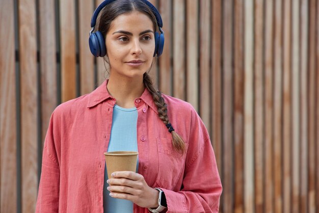 Une jeune femme européenne séduisante avec une queue de cochon porte un casque stéréo écoute de la musique tient un café à emporter porte une chemise rose pose contre du bois donc regarde au loin Concept de personnes et de style de vie