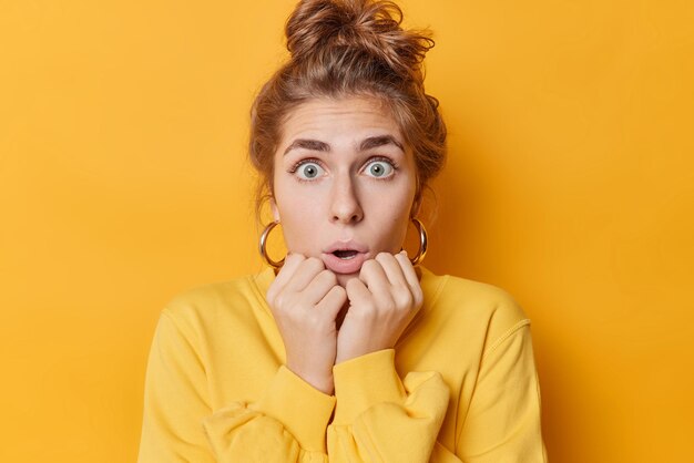 Une jeune femme européenne stupéfaite regarde les yeux sur écoute garde les mains sous le menton a les cheveux rassemblés en chignon porte un pull décontracté réagit aux nouvelles choquantes isolées sur fond jaune Concept d'émotions