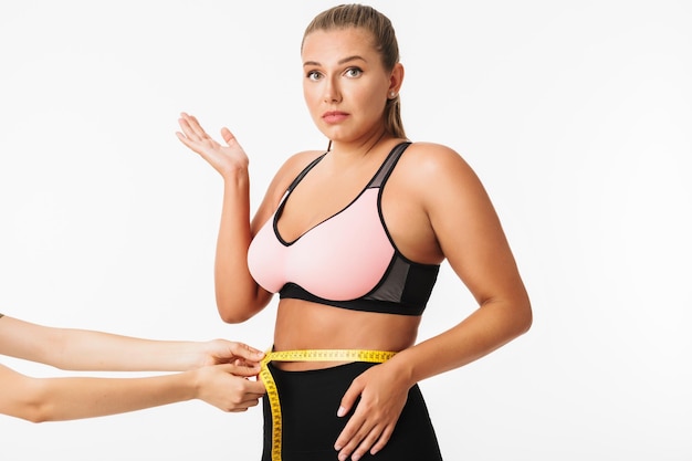 Jeune femme avec un excès de poids en haut sportif regardant étonnamment à huis clos tout en mesurant la taille sur fond blanc