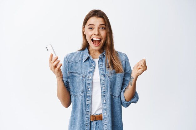 Une jeune femme excitée crie oui et souriante émerveillée, tenant un smartphone et regardant devant, gagnant en ligne, se tenant heureuse de sa réussite contre un mur blanc